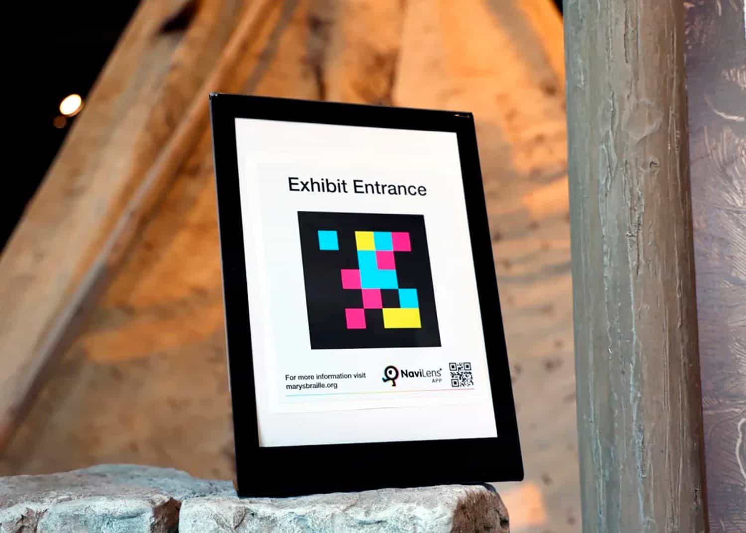 Black-framed “Exhibit Entrance” sign with a large NaviLens code, “NaviLens APP” logo and QR code, leaning on a stone wall inside the museum