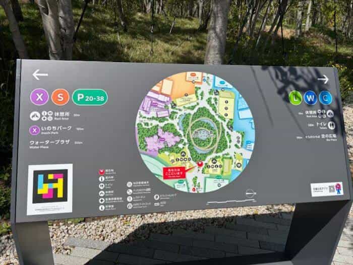 Site map sign with the circle of zones X, S, P, L, W, C labelling “Inochi Park / いのちパーク”, “Water Plaza / ウォータープラザ” and “Sora Plaza / 空の広場”, with a NaviLens code mounted in the bottom-left corner