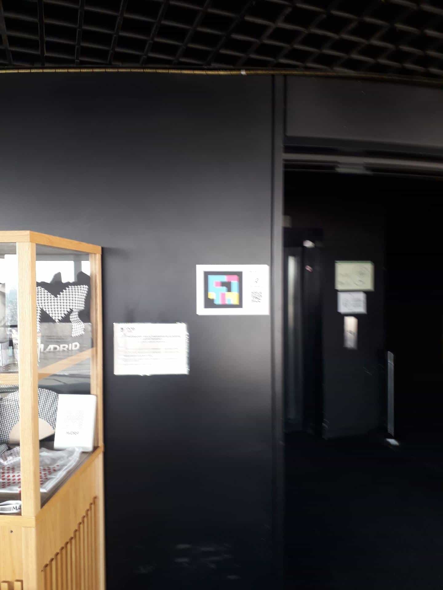 Lobby of the Faro de Moncloa viewpoint: on the black wall, a sign announces the NaviLens codes on the information railing, next to a display case with “Madrid” merchandising and the entrance to the panoramic lift