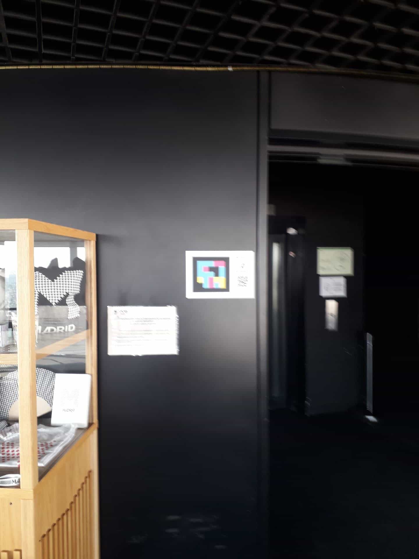 Atrio del belvedere del Faro de Moncloa: sulla parete nera un cartello annuncia i codici NaviLens del parapetto informativo, accanto a una vetrina con il merchandising «Madrid» e all’ingresso dell’ascensore panoramico