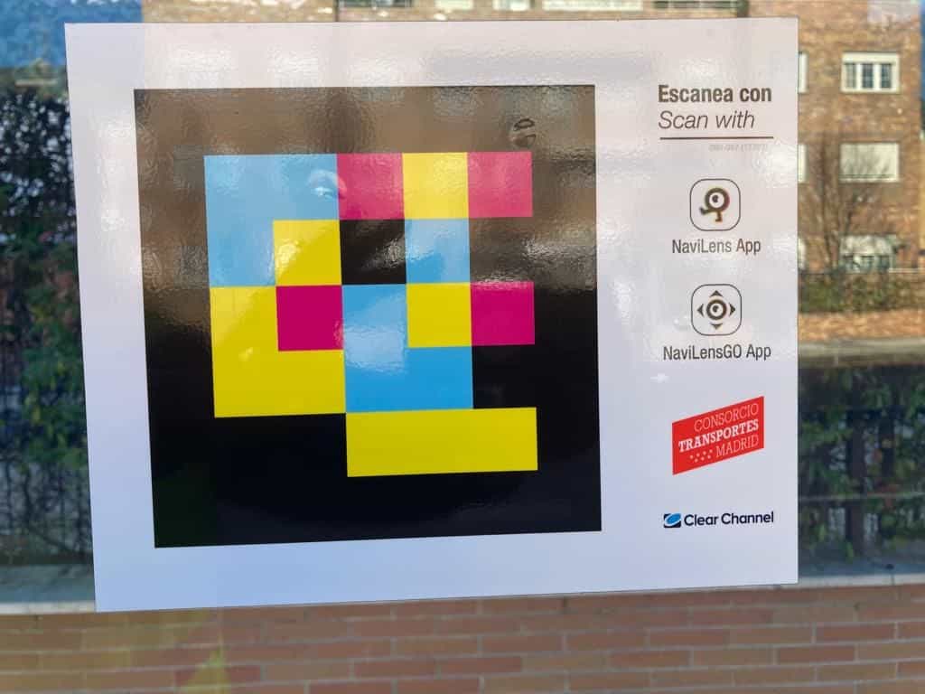 Cartel «Escanea con / Scan with» con un código NaviLens cuadrado a colores, iconos de las apps NaviLens y NaviLens GO, logo del CRTM y logo de Clear Channel pegado sobre el cristal de una marquesina
