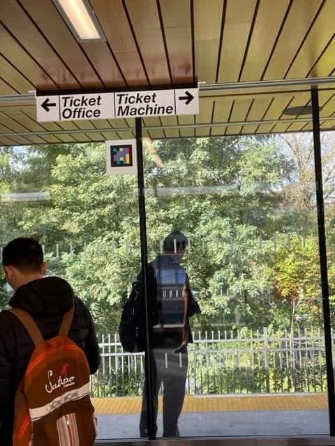 Ceiling-suspended sign with two arrows «← Ticket Office» and «Ticket Machine →»; below the sign, a NaviLens code; in the background, a backpacked traveller looking towards the tracks