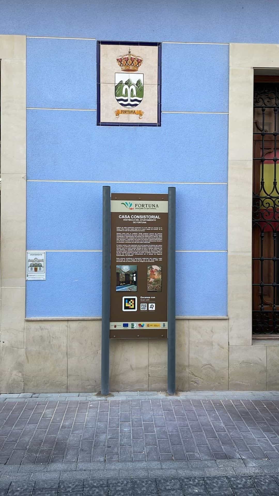 Fachada azul de la Casa Consistorial de Fortuna con el escudo municipal en cerámica y, debajo, un tótem marrón con la marca «Fortuna · Descubre lo que somos», cartela de la Casa Consistorial, código NaviLens cuadrado y placa ARASAAC con el pictograma «Ayuntamiento»