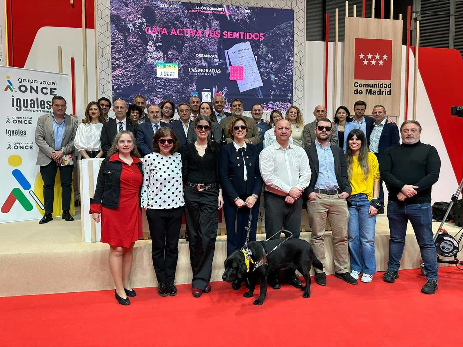 Foto di gruppo della degustazione «Activa tus Sentidos» al Salón Gourmets: un gruppo numeroso posa sulla pedana rossa dello stand della Comunità di Madrid, con un cane guida labrador nero della ONCE in primo piano e uno schermo in fondo con il manifesto dell’evento, organizzato da Grupo Social ONCE, Las Moradas de San Martín e Hostelería Madrid