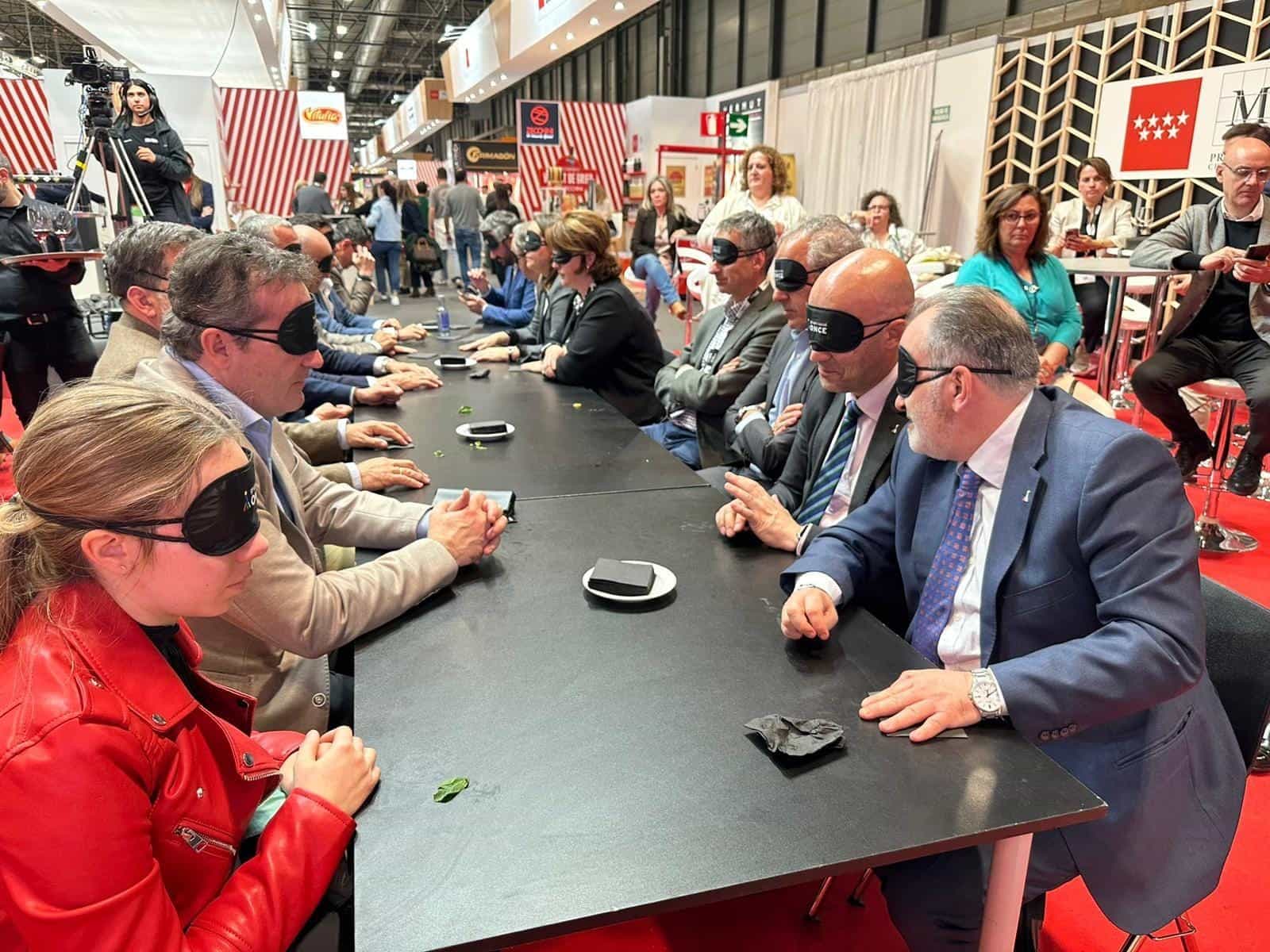 Tavolo lungo del Salón Gourmets a IFEMA durante la degustazione «Activa tus Sentidos» di Decanta Madrid: partecipanti con mascherine nere del Grupo Social ONCE seduti ai due lati, con piattini e briciole sul tavolo, in un corridoio fiancheggiato dagli stand della fiera