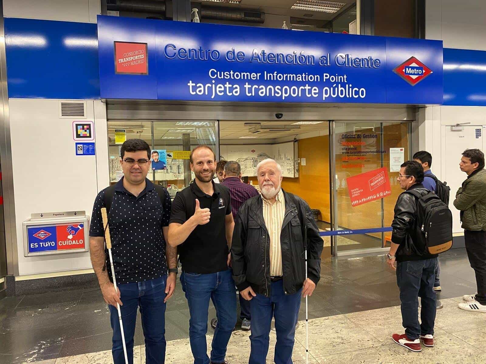 Metro de Madrid Customer Information Point at Nuevos Ministerios: blue corporate sign with the Metro red diamond and the Madrid Transport Consortium logo; on the metal side of the entrance, a square NaviLens code stuck at traveller's height. In front, two blind users with canes pose with a NaviLens technician