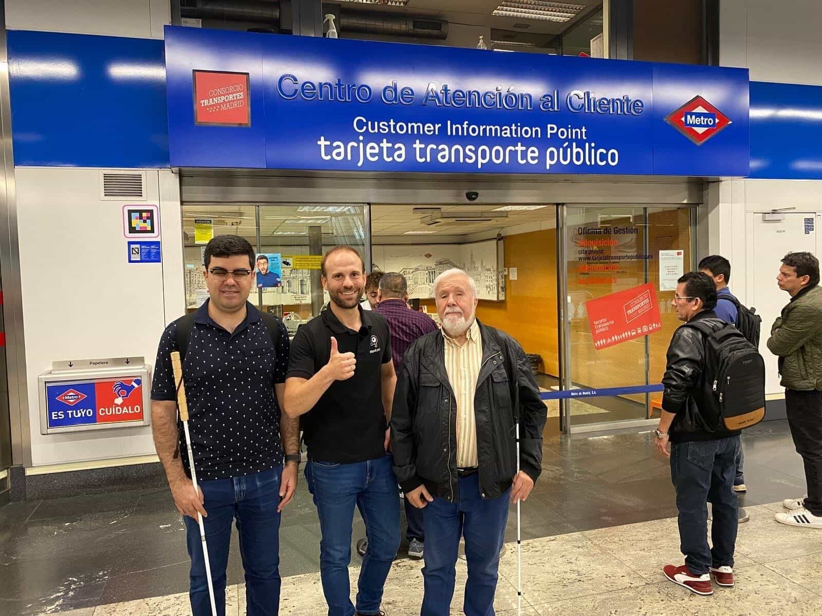 Kundenservice-Punkt von Metro de Madrid in Nuevos Ministerios: blaues Firmenschild mit roter Metro-Raute und Logo des Madrider Verkehrsverbunds; an der Metallseite des Eingangs ein quadratischer NaviLens-Code auf Fahrgasthöhe. Davor posieren zwei blinde Nutzer mit Stock und ein NaviLens-Techniker