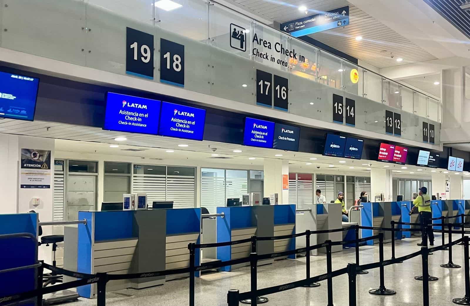 Innerer Check-in-Bereich des Flughafens Los Garzones in Montería: lange Schalter mit blauen Fluginformations-Monitoren, verchromte Absperrpfosten begrenzen die Warteschlangen, Passagiere mit Gepäck vor den Schaltern
