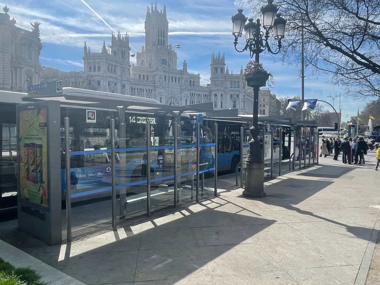 Veduta delle pensiline della EMT in Plaza de Cibeles, con un autobus blu della linea 14 diretto a Conde de Casal che esce dalla fermata; in fondo il Palacio de Comunicaciones (sede del Comune di Madrid). Sul frontale del tetto della pensilina, un codice NaviLens quadrato a colori