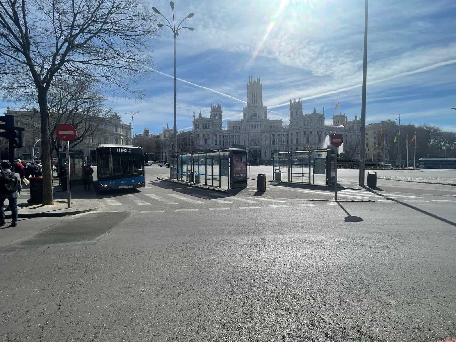 Plaza de Cibeles con diverse pensiline della EMT allineate sullo spartitraffico, un autobus blu n. 3262 in sosta e, in fondo, il Palacio de Comunicaciones sotto un cielo sereno: una delle fermate più frequentate di Madrid, integrata nella rete NaviLens