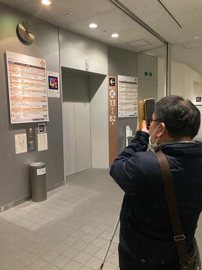 Civic Center のエレベーター横で、白杖を持つ視覚障害のある人がスマートフォンで NaviLens コードを読み取り、音声で情報を聞いている