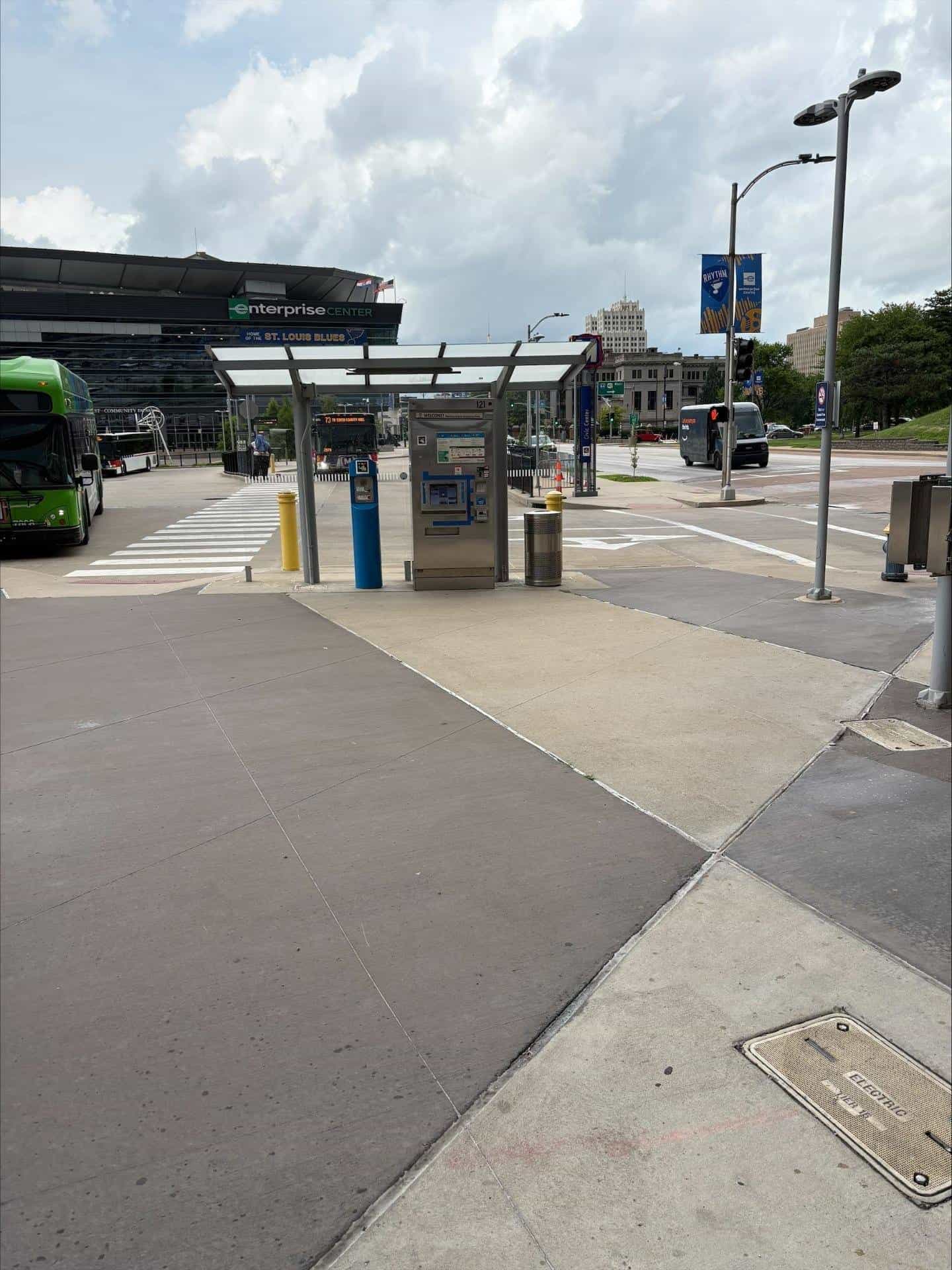 Exterior view of the Civic Center Transit Center in St. Louis: a flat white canopy on a grey metal frame covering a stainless steel ticket vending machine (TVM) with a blue "WELCOME" sign and a NaviLens code in yellow, pink, blue and black squares, next to a blue assistance post; in the background, the black facade of the Enterprise Center with the green sign and blue "HOME OF THE ST. LOUIS BLUES" stripe, two Metro buses (routes 73 South County Mall and 10), a white Amazon electric van and the downtown skyline under a cloudy sky