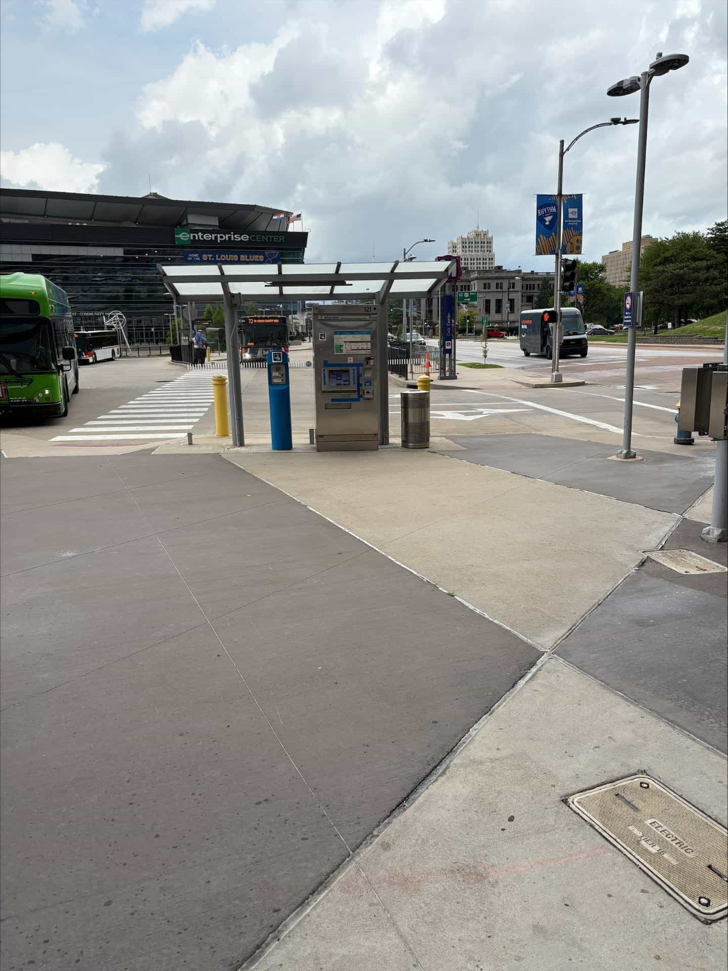 Außenansicht des Civic Center Transit Center in St. Louis: flaches weißes Vordach auf grauem Stahlrahmen über einem Edelstahl-Ticketautomaten mit blauem „WELCOME"-Schild und NaviLens-Code, daneben ein blauer Assistance-Pfosten; im Hintergrund die schwarze Fassade des Enterprise Center mit dem grünen Schriftzug und dem blauen Streifen „HOME OF THE ST. LOUIS BLUES", zwei grün-weiße Metro-Busse, ein weißer Amazon-Elektrolieferwagen und die Downtown-Skyline unter bewölktem Himmel