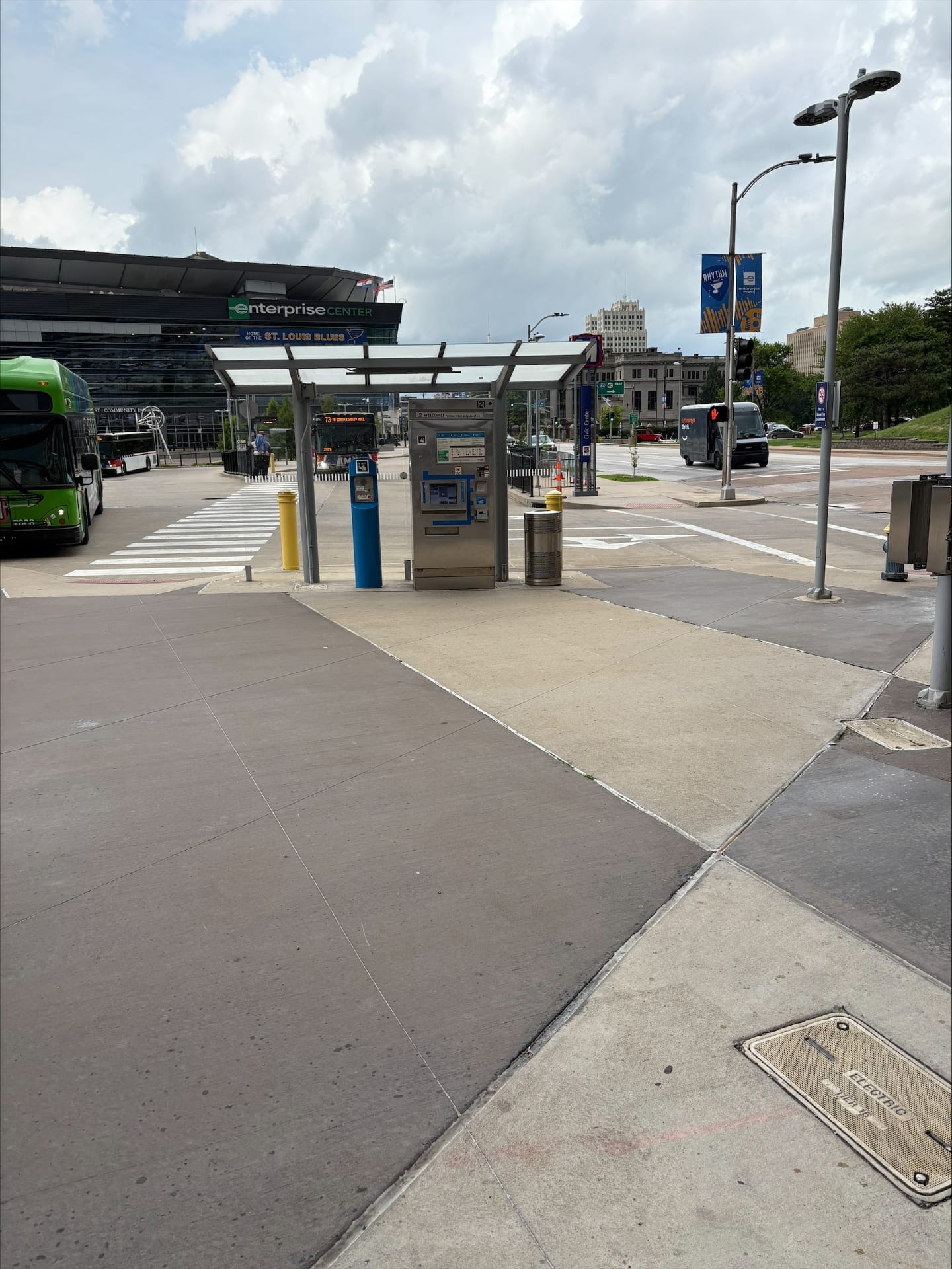 Exterior view of the Civic Center Transit Center in St. Louis: a flat white canopy on a grey metal frame covering a stainless steel ticket vending machine (TVM) with a blue "WELCOME" sign and a NaviLens code in yellow, pink, blue and black squares, next to a blue assistance post; in the background, the black facade of the Enterprise Center with the green sign and blue "HOME OF THE ST. LOUIS BLUES" stripe, two Metro buses (routes 73 South County Mall and 10), a white Amazon electric van and the downtown skyline under a cloudy sky
