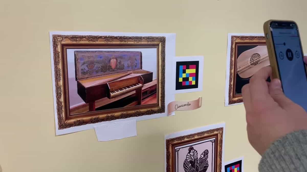 Cuadro del clavicordio con marco dorado, cartela en cinta marrón, código NaviLens al lado y etiqueta blanca con relieve táctil en braille; un usuario sostiene el móvil que reproduce el audio descriptivo