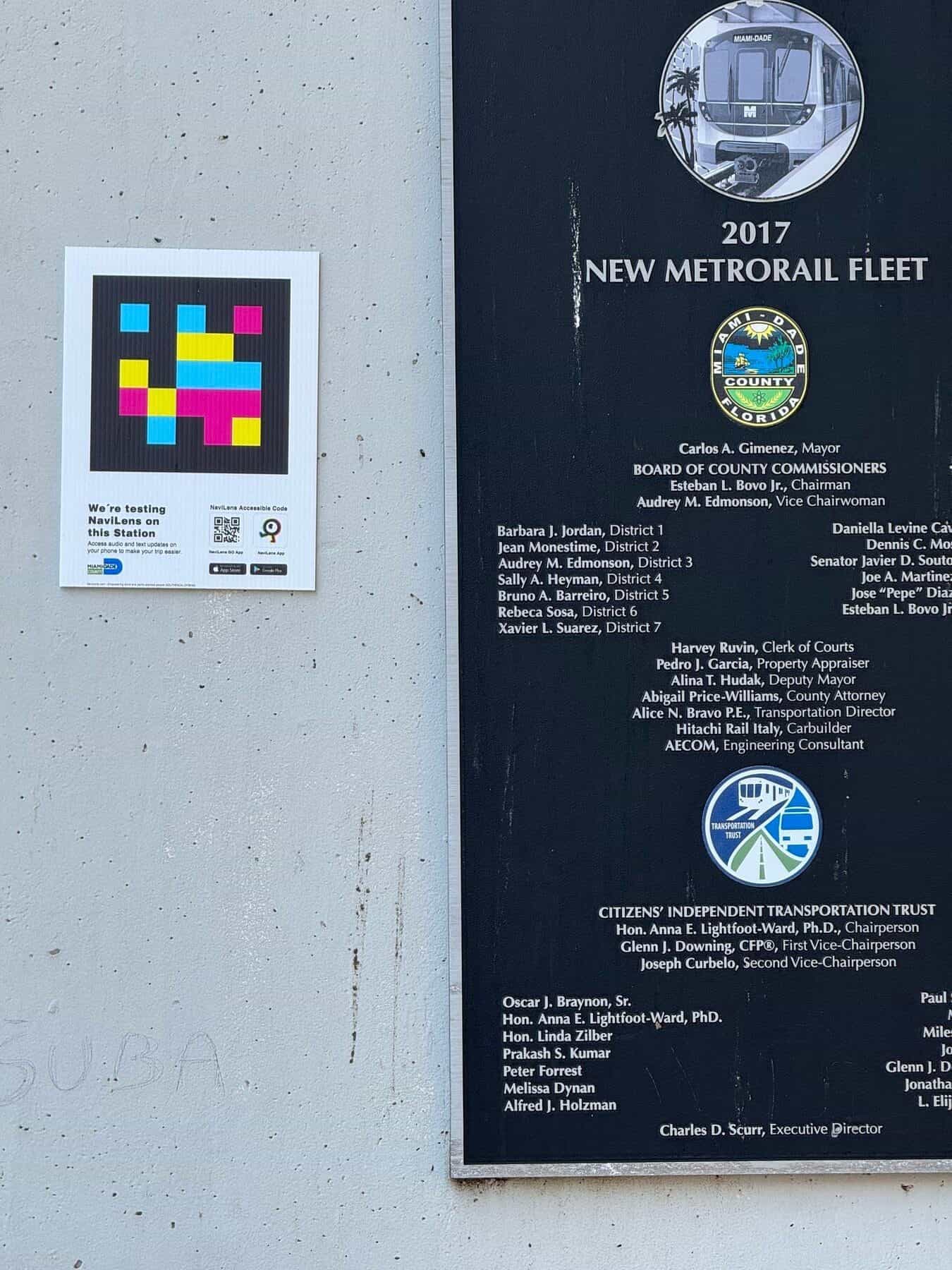 Dettaglio della targa commemorativa «2017 New Metrorail Fleet» con il sigillo della contea di Miami-Dade e l’elenco delle cariche pubbliche, accanto a un cartello NaviLens quadrato con la scritta «We’re testing NaviLens on this Station» e i QR per scaricare le app NaviLens e NaviLens GO