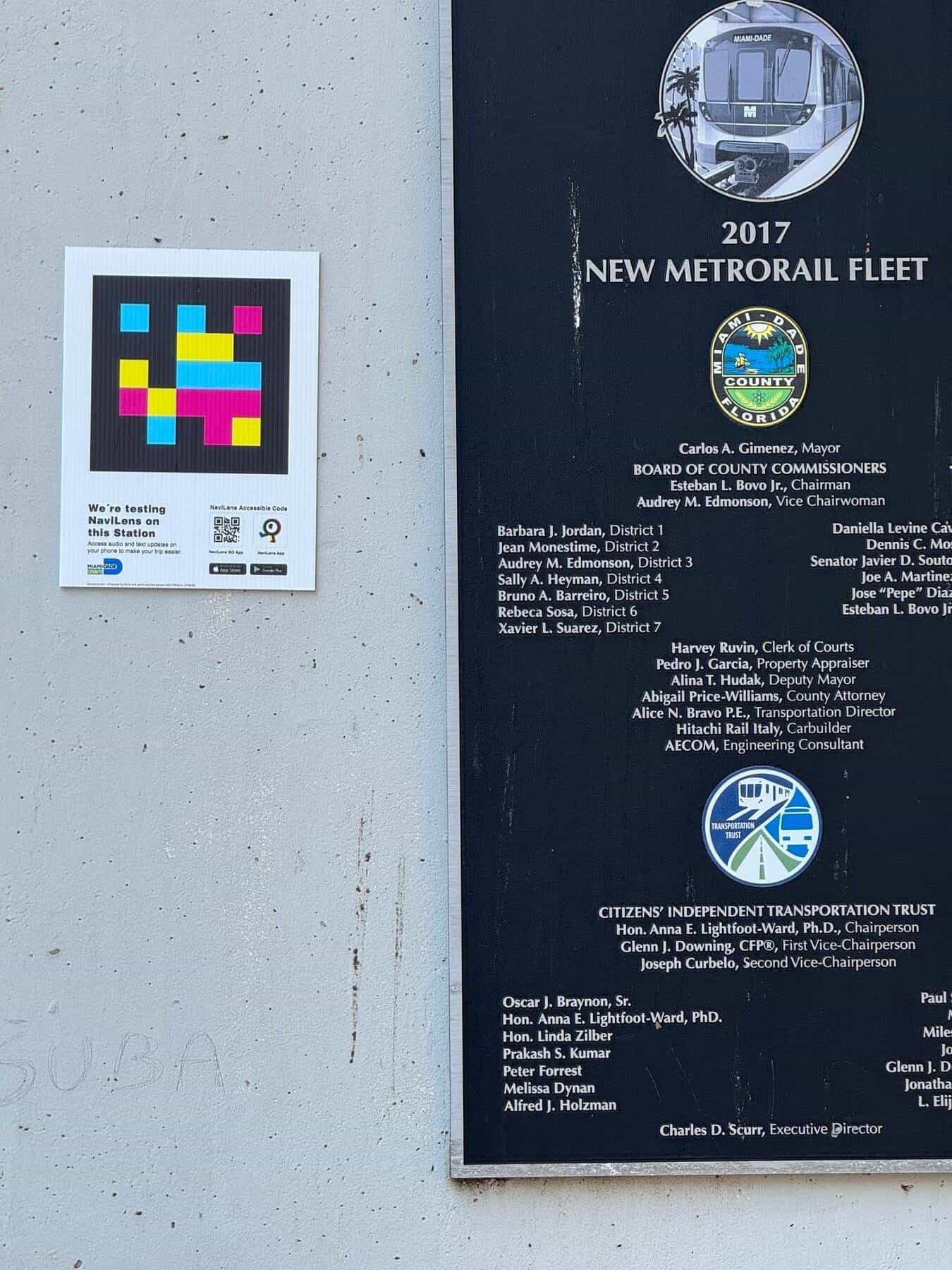 Close-up of the “2017 New Metrorail Fleet” commemorative plaque with the Miami-Dade County seal and the list of public officials, next to a square NaviLens sign reading “We're testing NaviLens on this Station” with QR codes to download the NaviLens and NaviLens GO apps