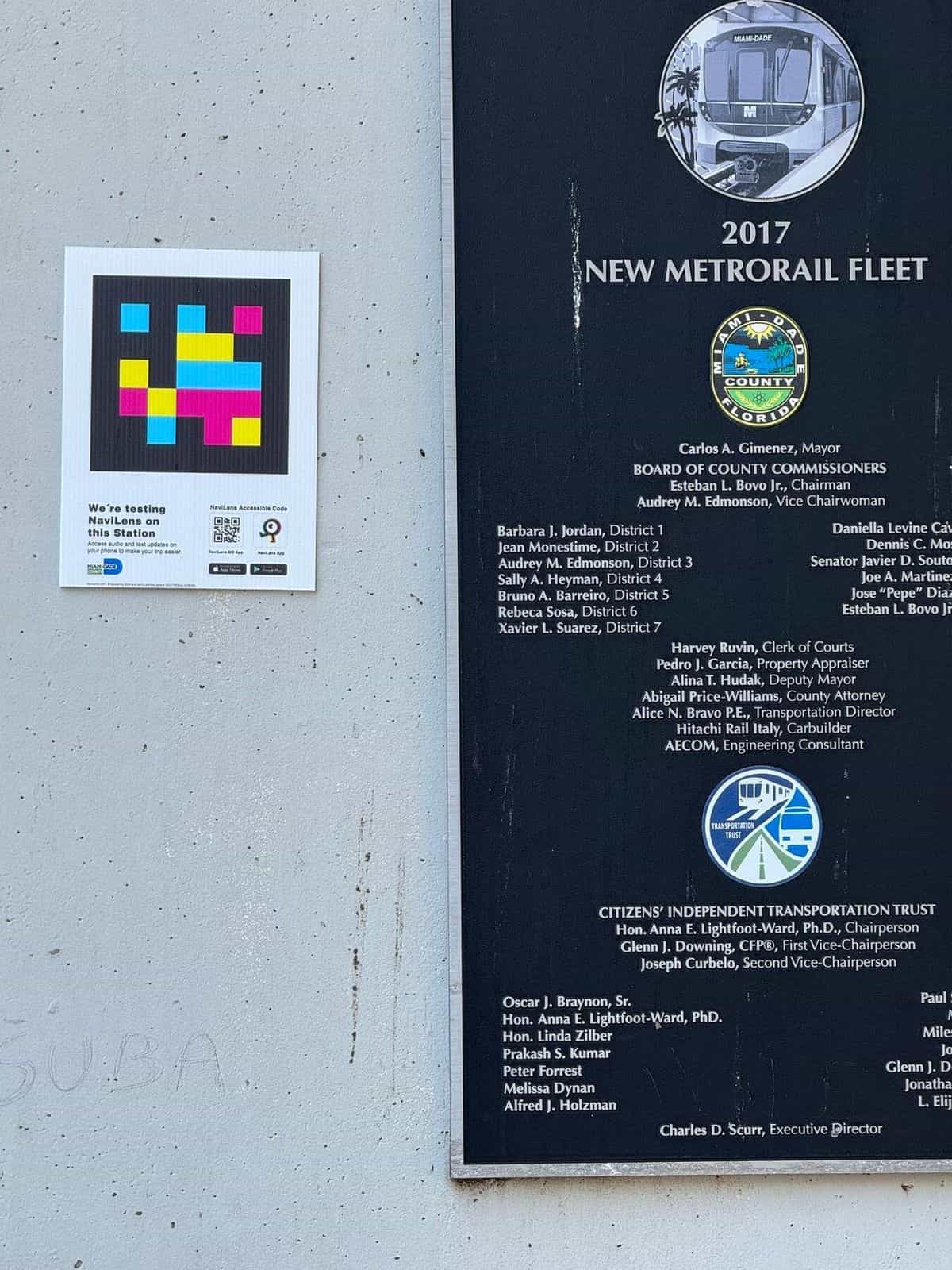 Detalle de la placa conmemorativa «2017 New Metrorail Fleet» con el sello del condado de Miami-Dade y la lista de cargos públicos, junto a un cartel NaviLens cuadrado con la leyenda «We're testing NaviLens on this Station» y los QR de descarga de las apps NaviLens y NaviLens GO