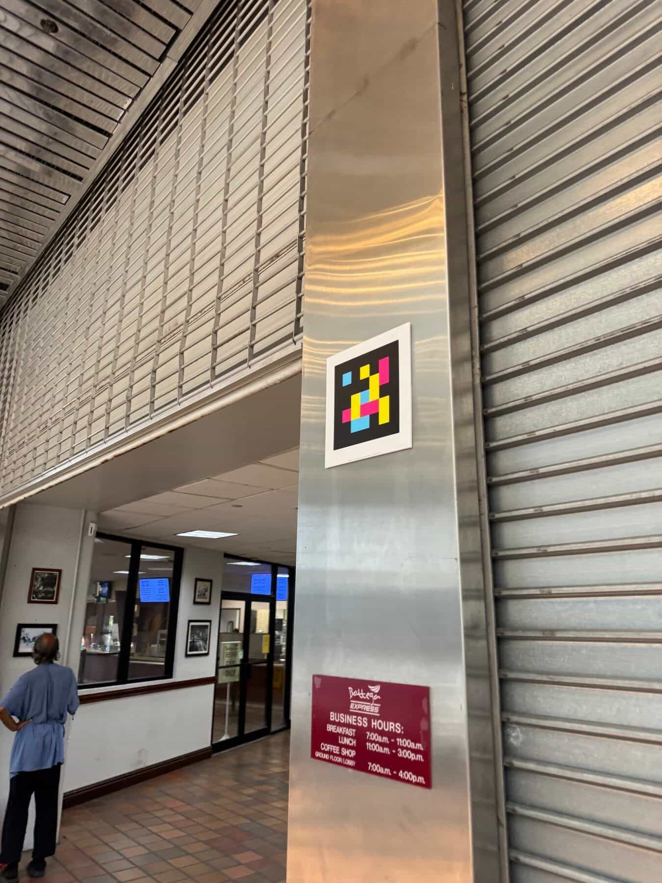 Atrio interno di Government Center con soffitto a doghe di legno scuro, colonna metallica e un codice NaviLens con cornice bianca fissato a mezza altezza, sopra il cartello degli orari del Bottega Express del Ground Floor / Lobby