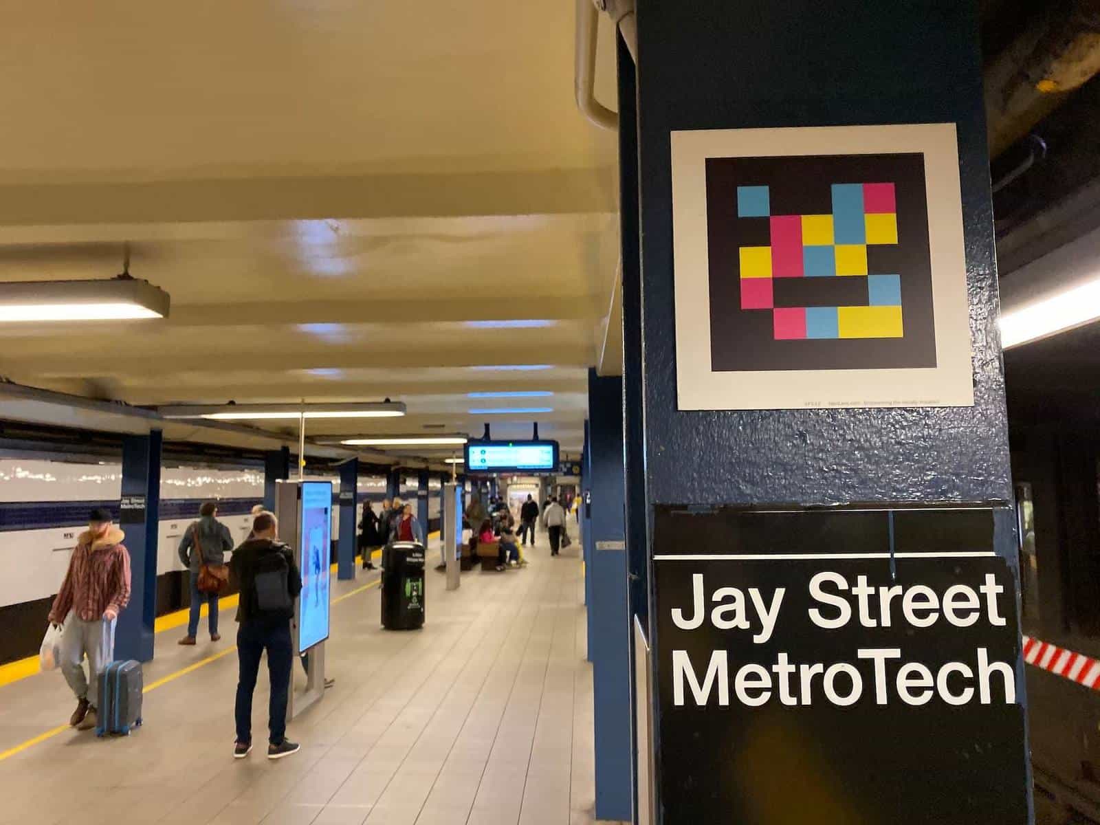 Side view of the Jay St-MetroTech platform with riders walking with suitcases and checking the digital panels. In the foreground, a blue column with the «Jay Street MetroTech» sign and a NaviLens code captioned «NaviLens.com — Empowering the visually impaired»