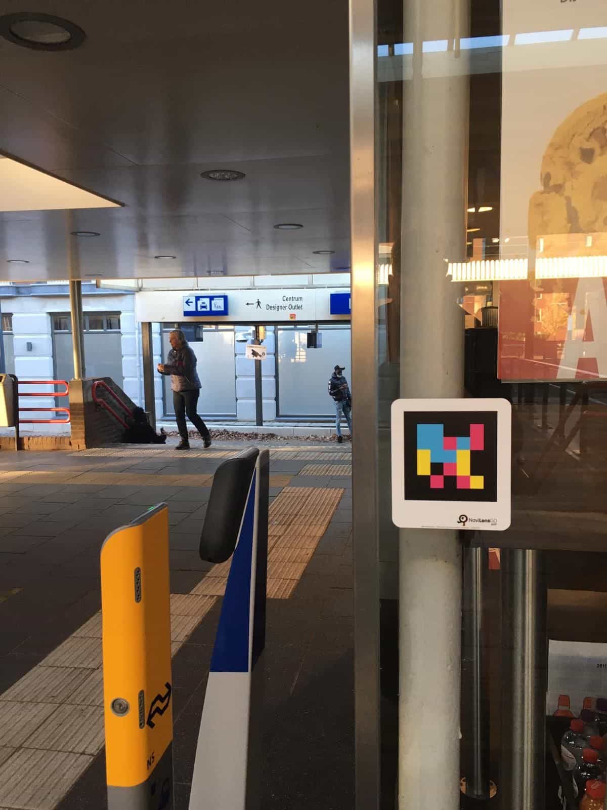 Codice NaviLens applicato su una colonna di vetro all’accesso della stazione degli autobus, con la segnaletica azzurra Centrum / Designer Outlet in fondo