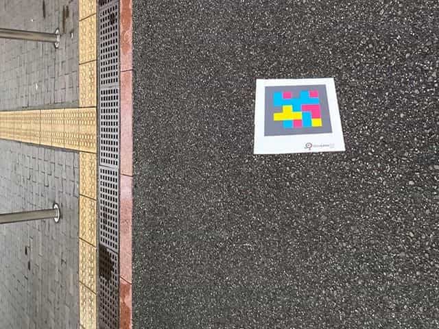 ポートアイランドのアスファルト上、排水グレーチングと美術館へ続く黄色い点字ブロックの隣に貼られたNaviLens GOタグ