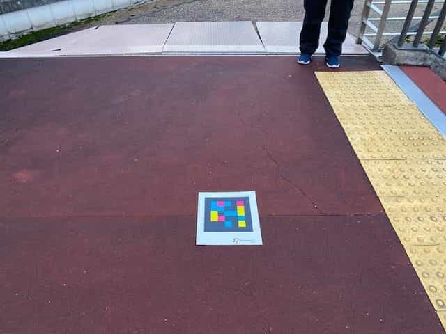 黄色い点字ブロックの縁と青いスニーカーを履いた参加者の足元の隣、赤い連続コンクリートの広場に置かれたNaviLens GOタグ