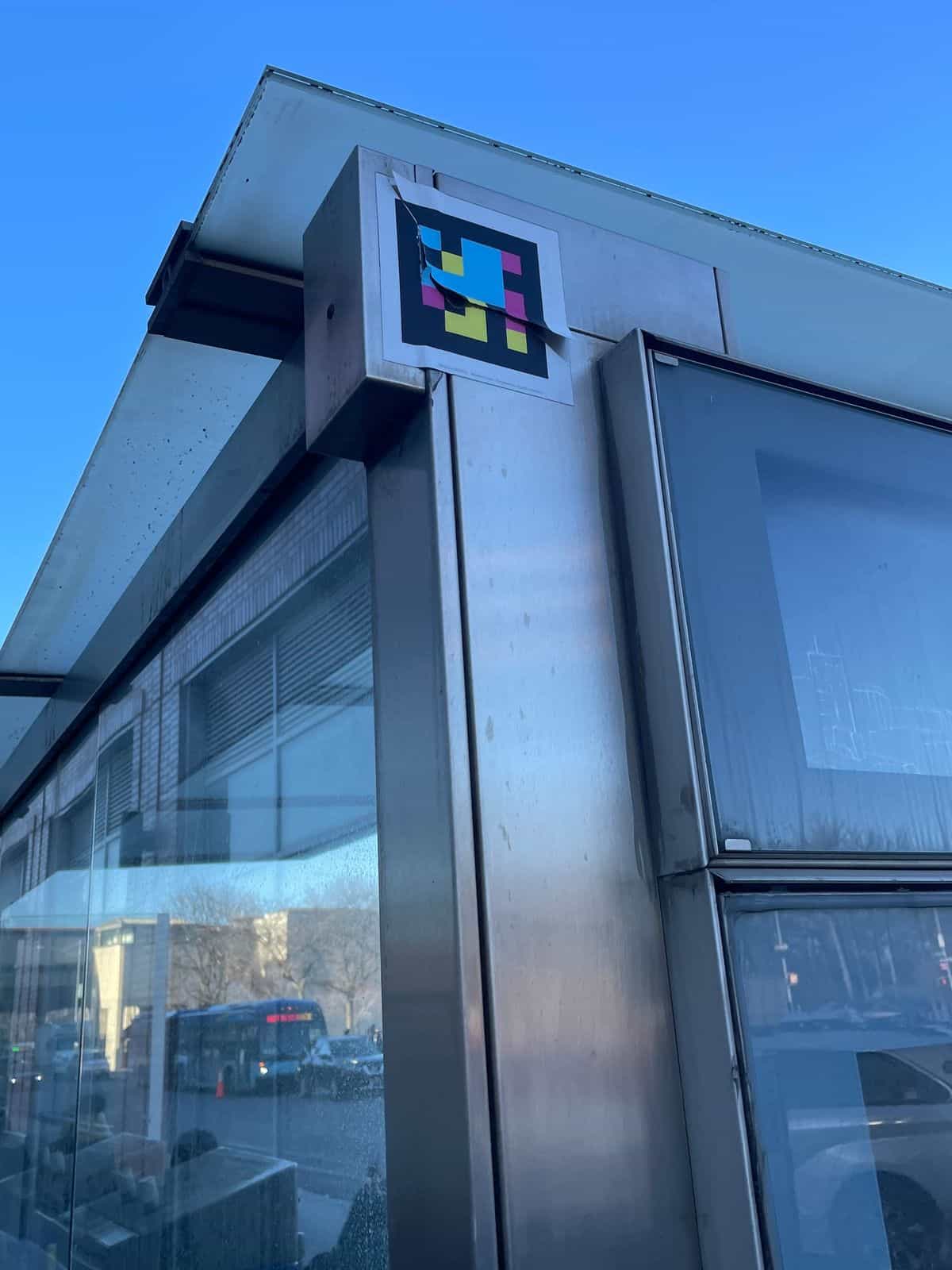 Detalle cercano de un código NaviLens pegado en el lateral superior de una marquesina del Bronx, mostrando su colocación sobre acero inoxidable y vidrio