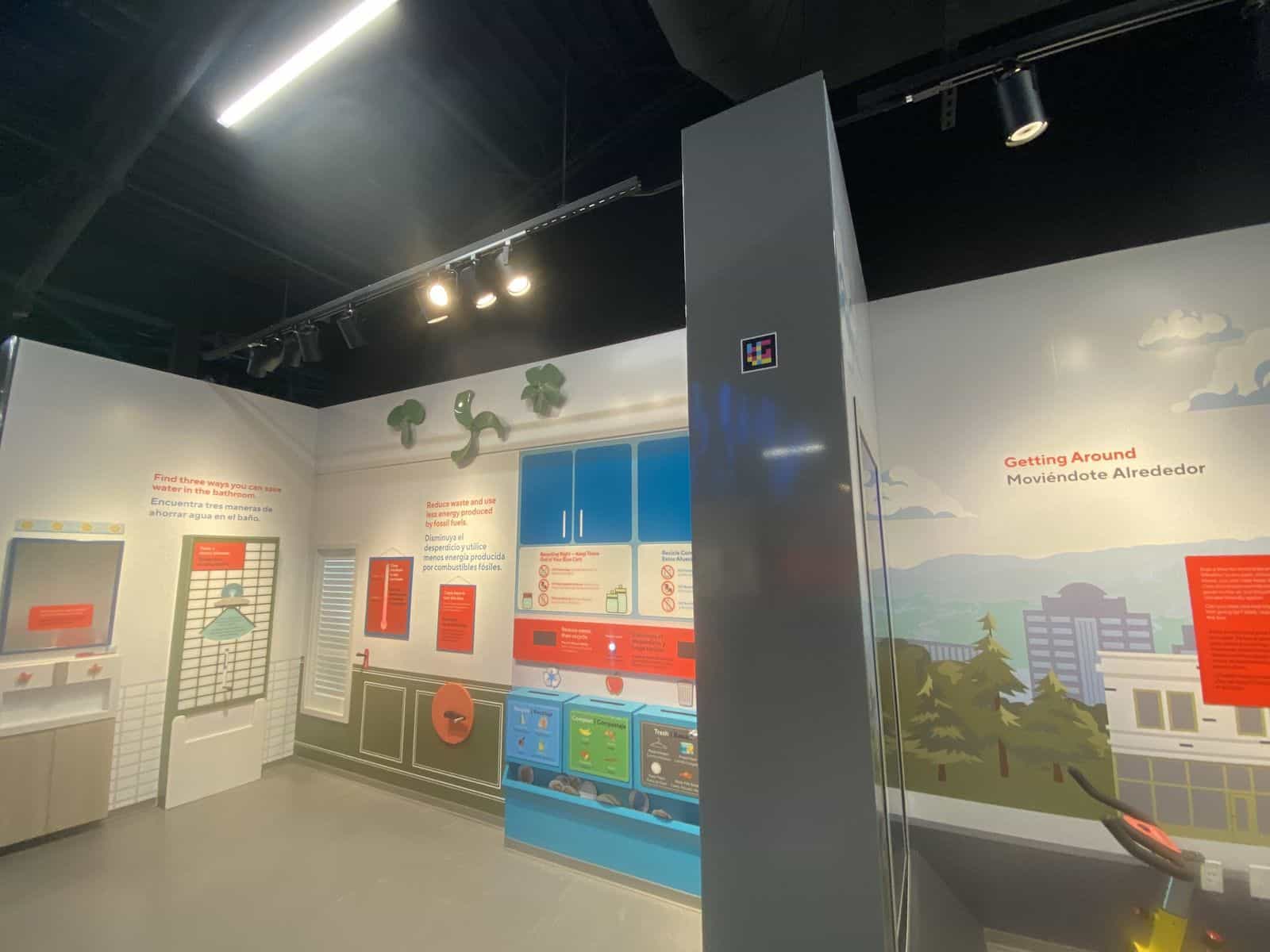 Säule in der Ausstellungshalle von The WaterShed mit einem NaviLens-Code auf Augenhöhe; dahinter das Wandbild „Getting Around / Moviéndote Alrededor“ und die Station zur Mülltrennung im Haushalt