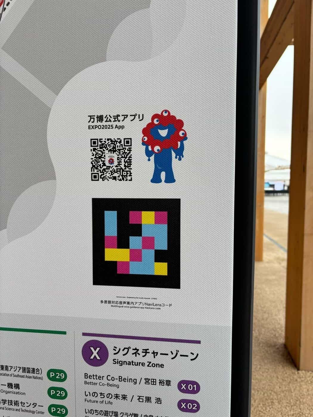 黒地にシアン・マゼンタ・イエローのNaviLensコードのマクロ詳細。「多言語対応音声案内アプリ NaviLensコード · Multilingual voice guidance app NaviLens code」のシールとEXPO2025アプリQRコードに重なる万博公式マスコット