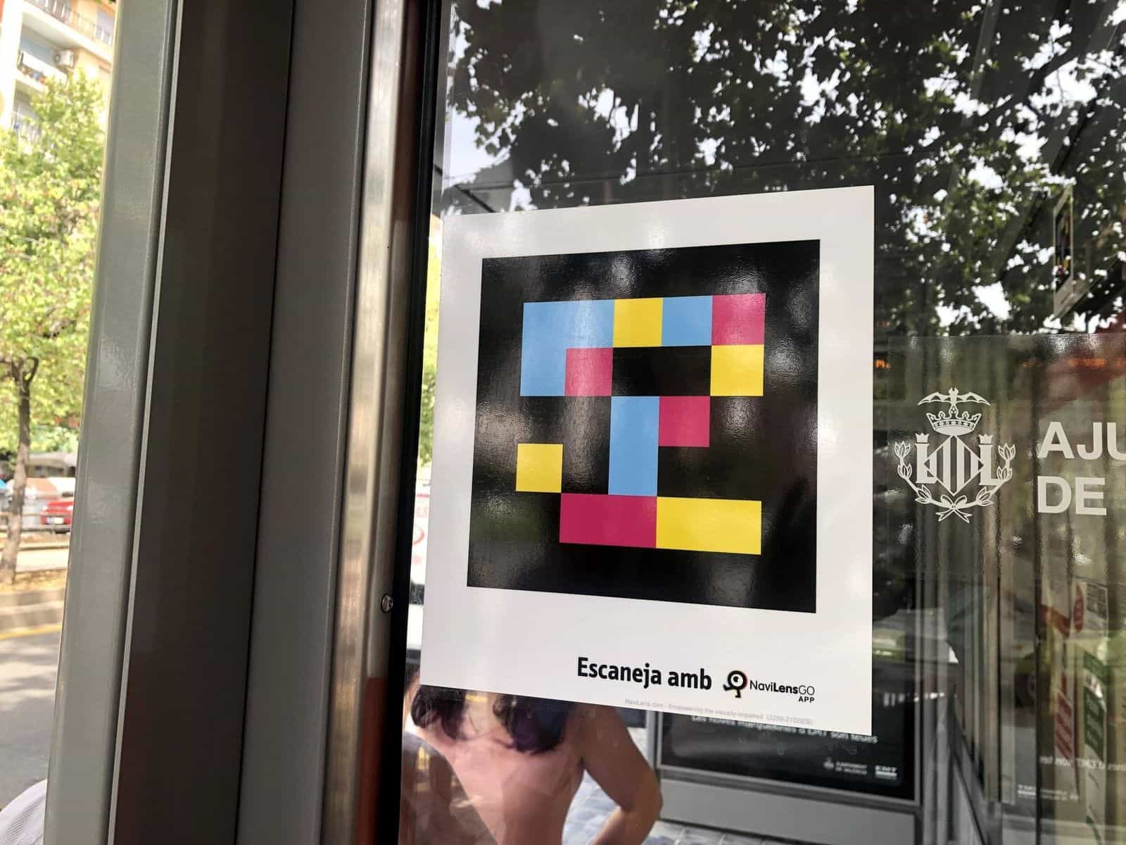 Dettaglio di un cartello applicato sul vetro della pensilina con un codice NaviLens quadrato a colori —quadrati blu, gialli e magenta su fondo nero— e sotto il testo «Escaneja amb NaviLens GO App»