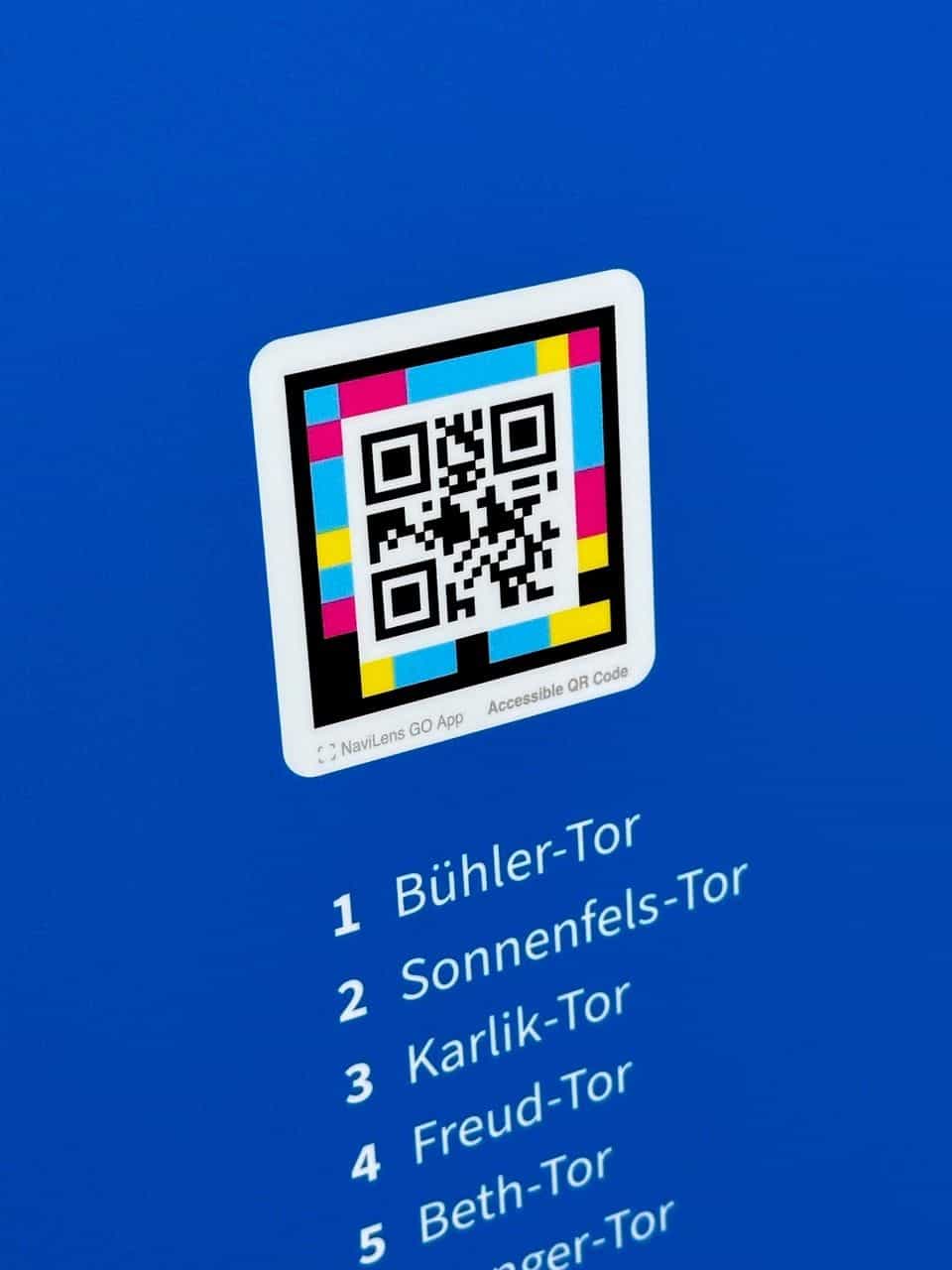 Nahaufnahme der neuen blauen Tafel der Tore der Erinnerung: quadratischer mehrfarbiger NaviLens-Code mit Marke „NaviLens GO App · Accessible QR Code“ und weiße typografische Liste der nummerierten Tore auf gesättigtem Universitätsblau