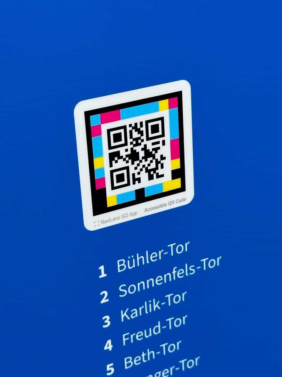 Close-up of the new blue Tore der Erinnerung panel: square multicolour NaviLens code with the “NaviLens GO App · Accessible QR Code” mark and a white typographic list of the numbered Tore on a saturated university-blue background