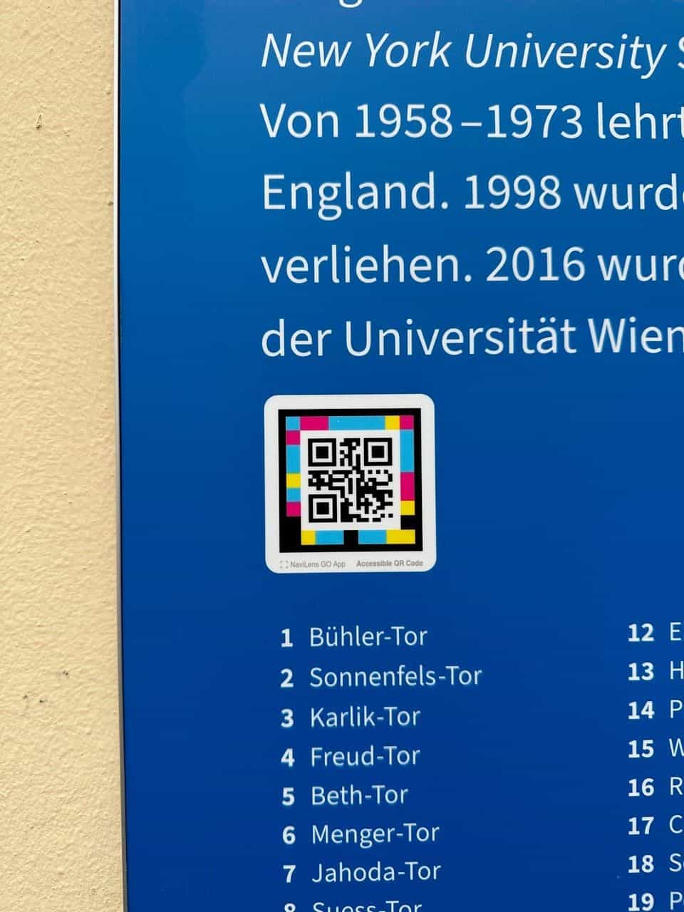 Detail der blauen Tafel der Tore der Erinnerung im Bereich „New York University“: weißer Text auf Blau, mehrfarbiger NaviLens-Code mit Marke „NaviLens GO App · Accessible QR Code“