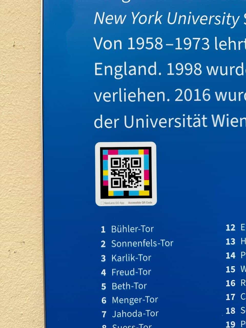 Close-up of the blue Tore der Erinnerung panel around the “New York University” section: white text on blue, a colour-coded NaviLens square with the “NaviLens GO App · Accessible QR Code” mark
