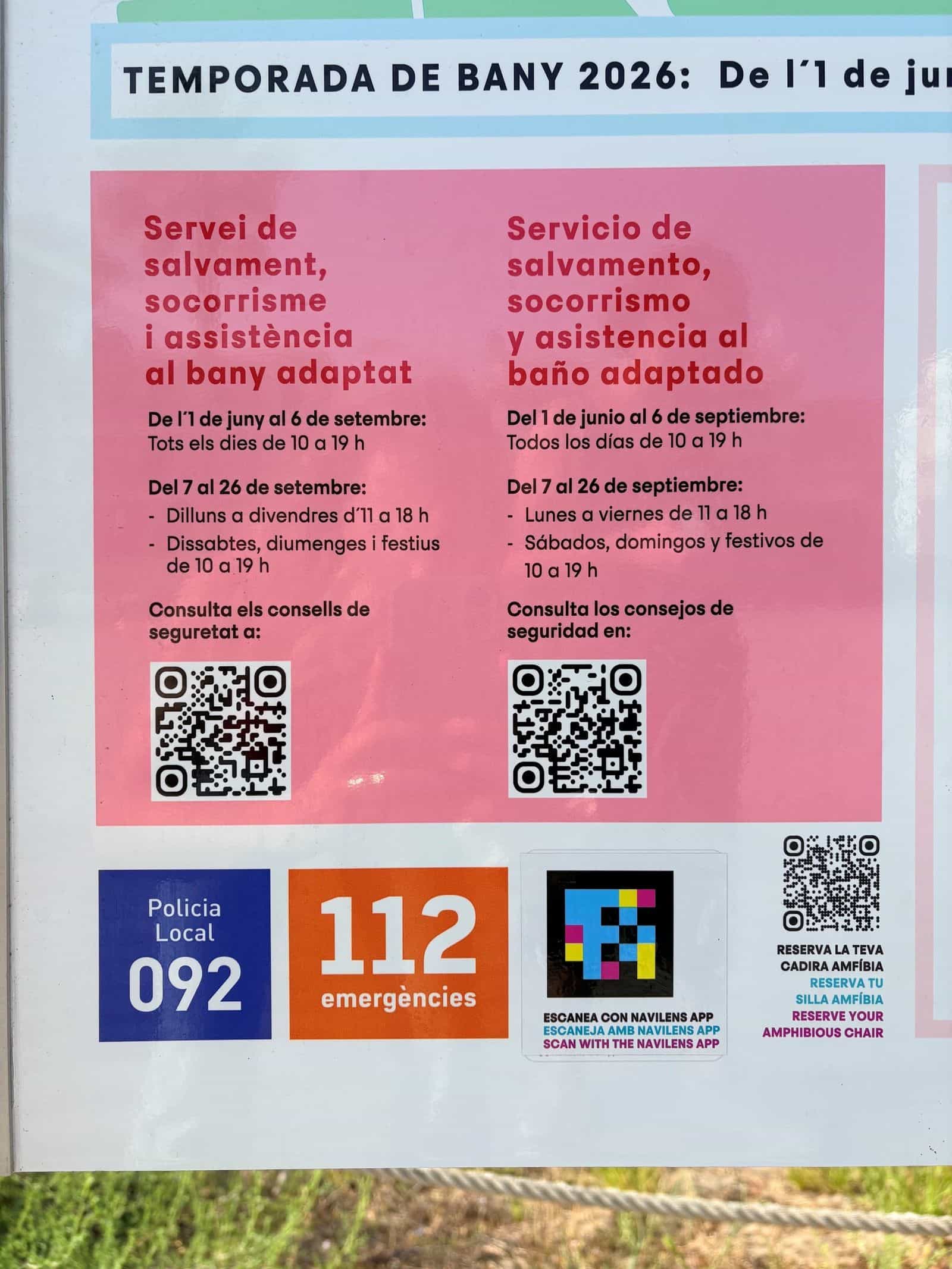Detail einer Strandtafel in Castelldefels: der Block „Servei de salvament, socorrisme i assistència al bany adaptat“ mit den Zeiten der Badesaison 2026 und seinen QR-Codes, darunter die 092 der Ortspolizei, die Notrufnummer 112 und der Aufkleber mit dem NaviLens-Code und dem Hinweis „ESCANEA CON NAVILENS APP“