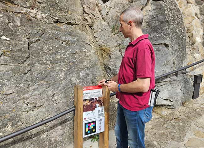 Ein Besucher liest mit der Hand die Tafel „Pas de la Foradada“ in Sant Miquel del Fai: auf Holzpfosten die Karte mit dem Reliefbild, der Block „Info“ und der NaviLens-Code aus farbigen Quadraten, montiert an der Felswand neben dem Metallhandlauf
