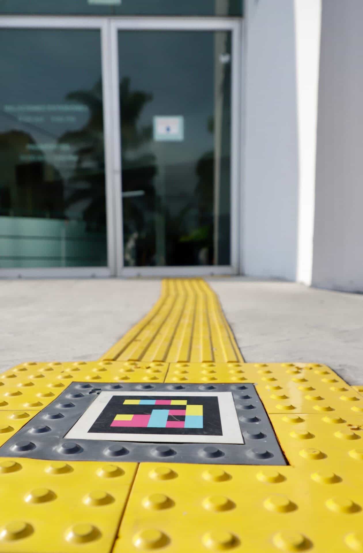NaviLens code embedded in the yellow tactile paving right in front of the glass door of the Zapopan Foreign Affairs Passport Office, with a continuous guiding strip towards the entrance