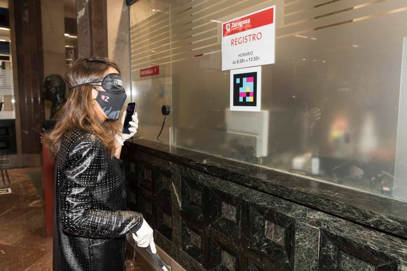 ONCE user with blindfold and cane reading with her mobile phone the NaviLens code stuck on the glass of Window 1 of the Zaragoza City Council Registry