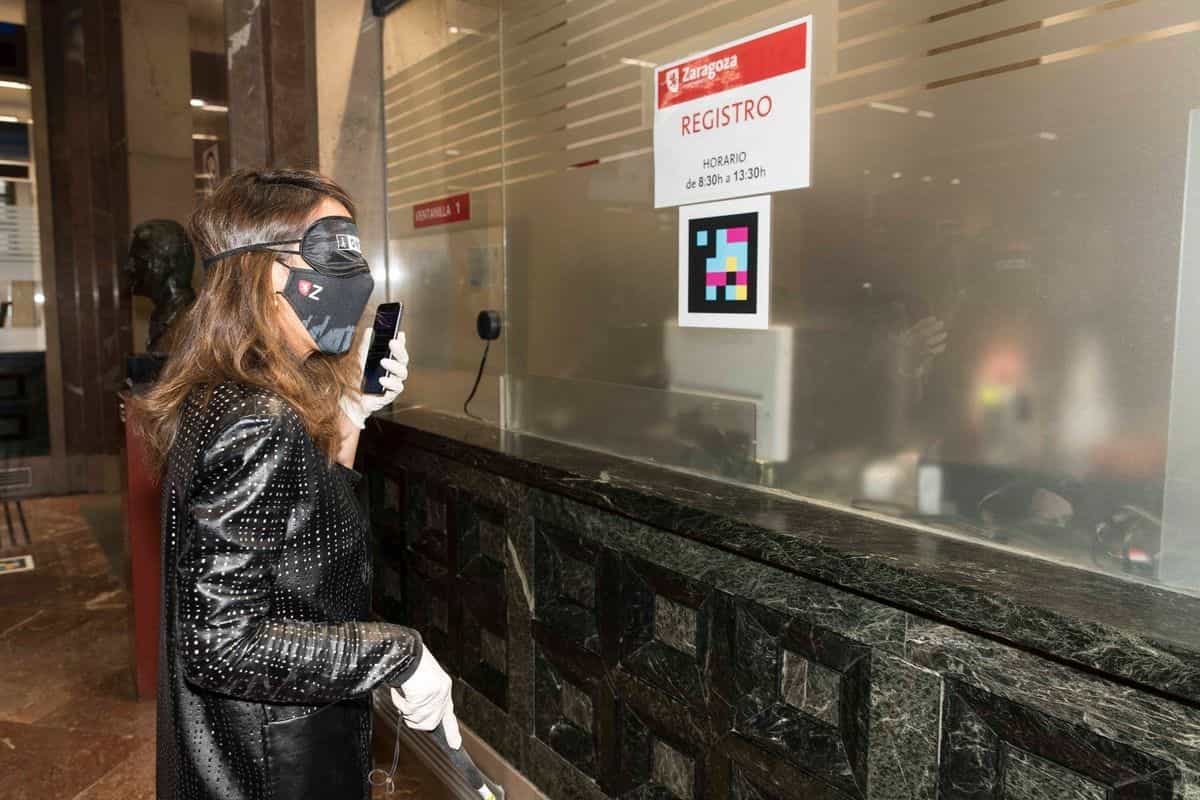 Usuaria de la ONCE con antifaz y bastón leyendo con su móvil el código NaviLens pegado en el cristal de la Ventanilla 1 de Registro del Ayuntamiento de Zaragoza