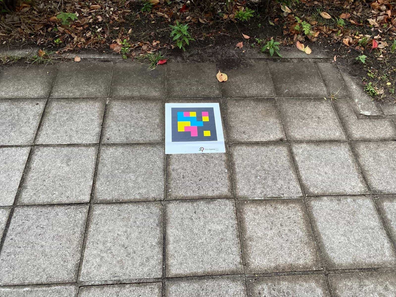 Large-format NaviLens GO tag on the garden paving of the Bandō Kagaku Kan, with fall leaves and vegetation in the background