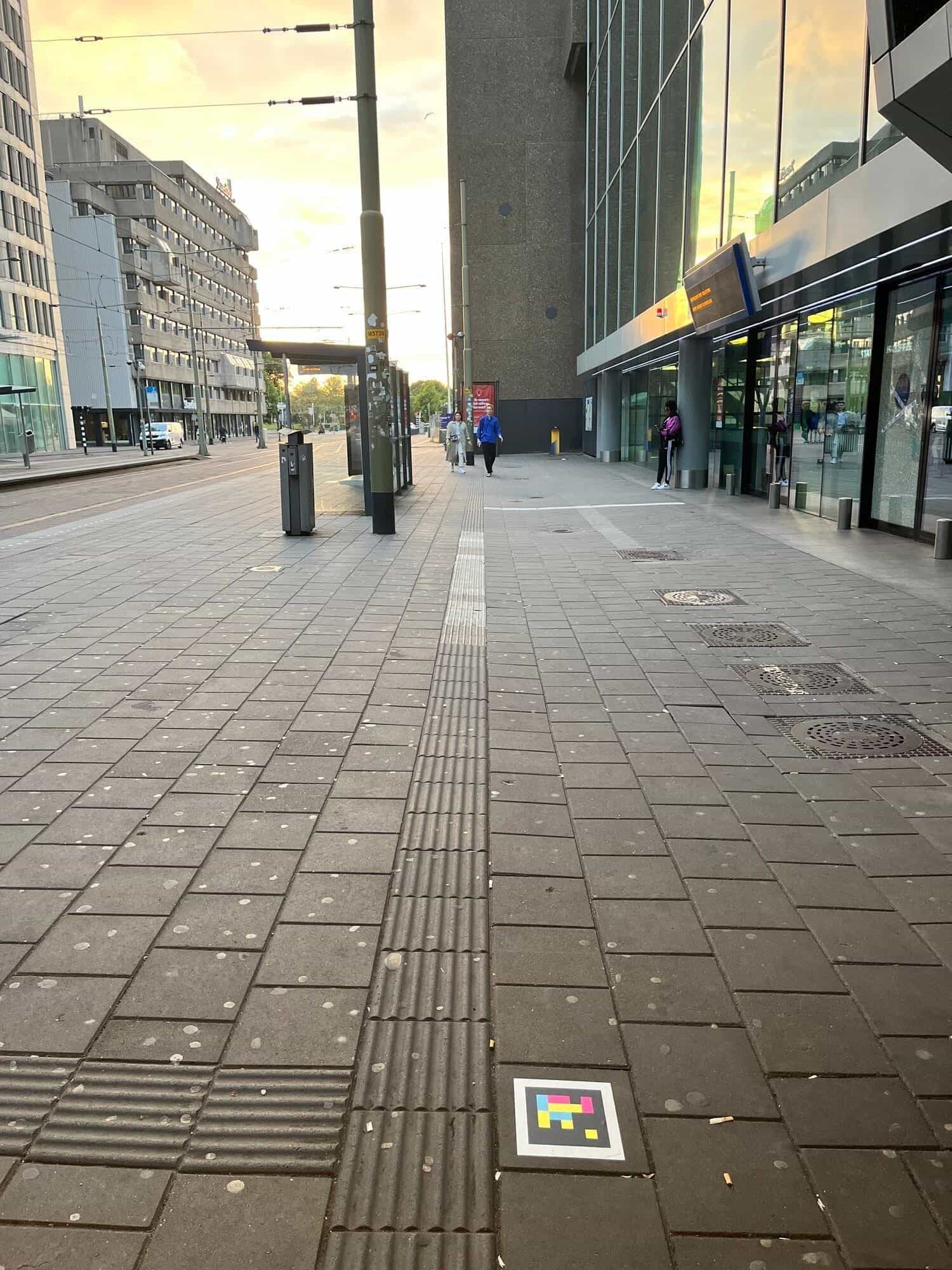 Accesso alla galleria commerciale di Den Haag Centraal con un codice NaviLens fissato alla parete sopra l’ingresso di Starbucks; il pannello LED di benvenuto mostra «Welkom op station Den Haag Centraal»