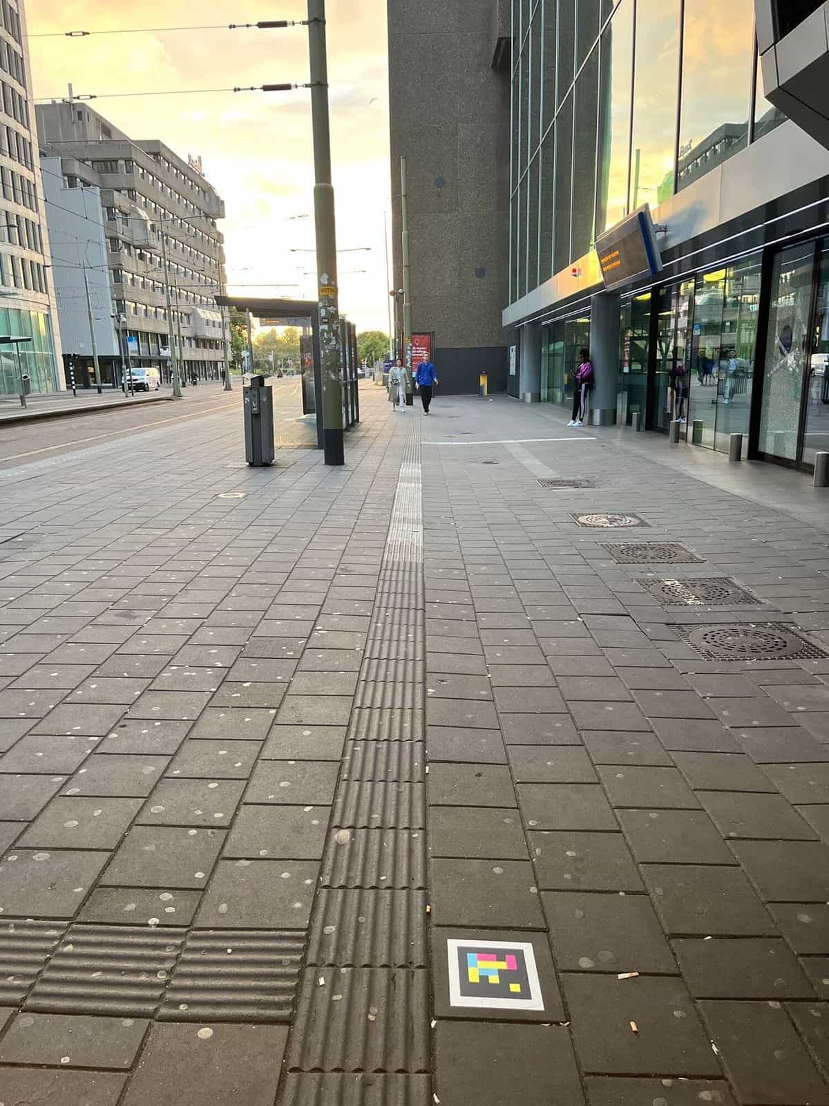 Acceso al hall comercial de Den Haag Centraal con un código NaviLens fijado en la pared sobre la entrada de Starbucks; el panel LED de bienvenida muestra «Welkom op station Den Haag Centraal»