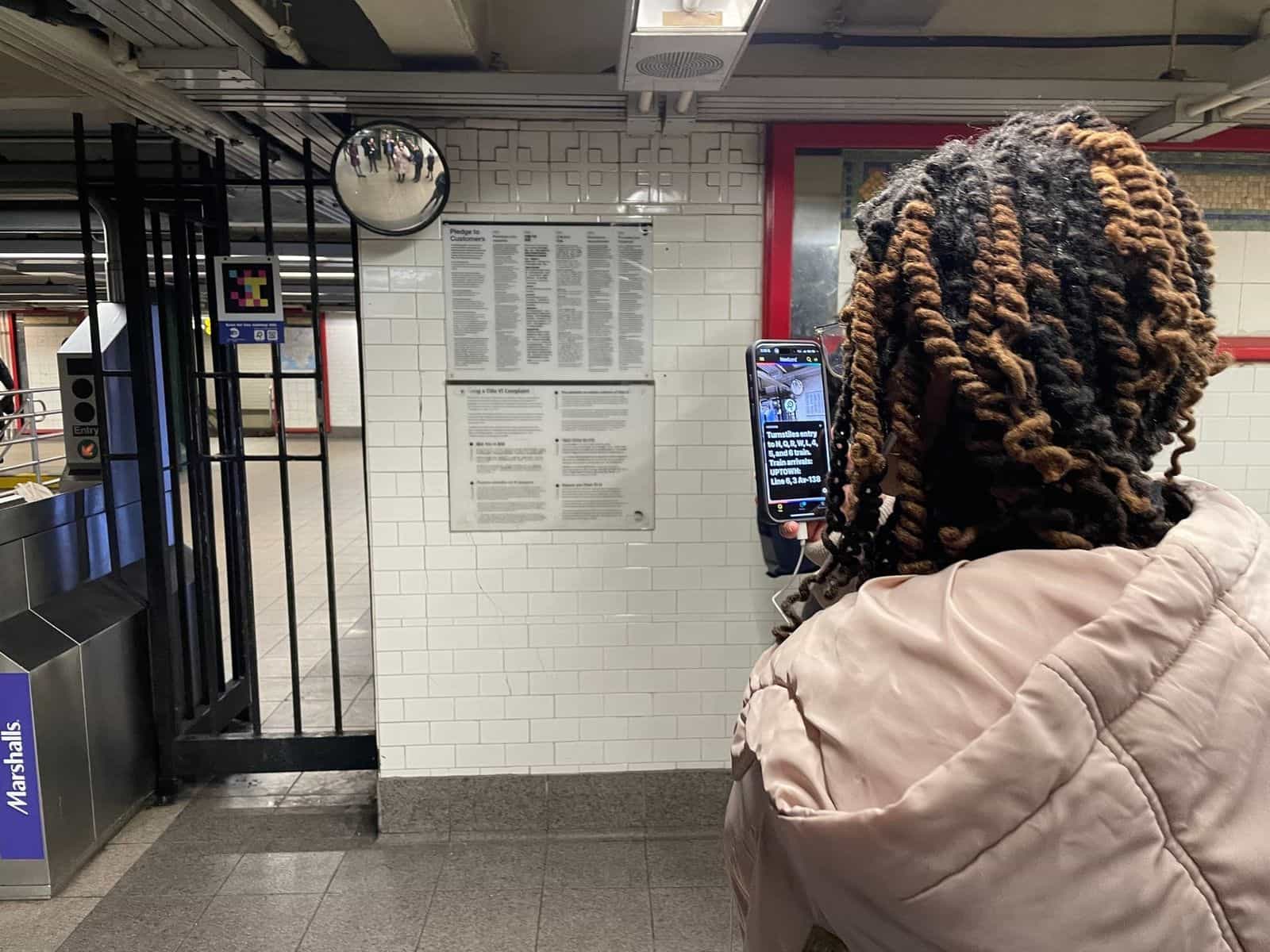 NaviLens code fixed next to the “Pledge to Customers” sign in the 14 St-Union Square concourse; in the foreground, a user holds the phone reading on screen “Turnstiles entry to N, Q, R, W, L, 4, 5 and 6 train. Train arrivals: UPTOWN: Line 6, 3 Av-138”