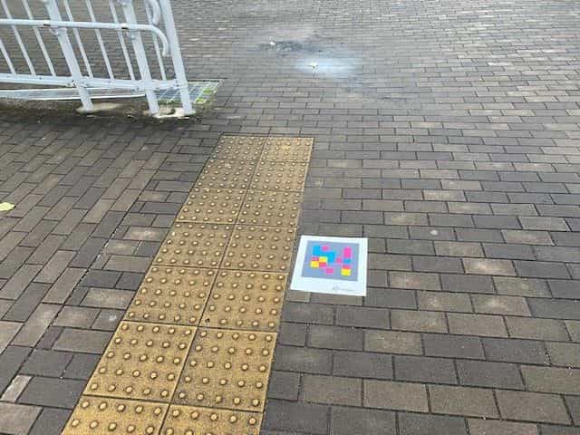 バンドー神戸青少年科学館のアクセススロープの白い手すり脇、黄色い点字ブロックの終端の石畳に貼られたNaviLens GOタグ