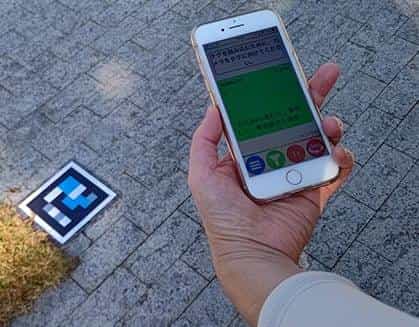 Hand holding an iPhone showing Japanese text while pointing at a cyan and magenta NaviLens code stuck on the museum's cobbled paving