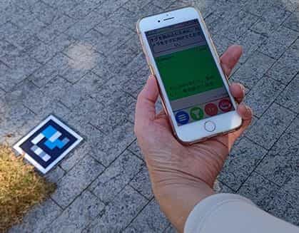 Hand hält ein iPhone mit japanischem Text und richtet es auf einen cyan-magentafarbenen NaviLens-Code auf dem Kopfsteinpflaster des Museums