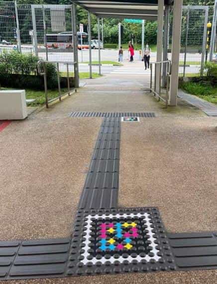 Outdoor covered crossing of the interchange with metal railings and greenery in the background: a grey T-shaped tactile guide line crosses the floor and ends on a square white tile with a large NaviLens code in coloured squares; in the background, a pedestrian crossing and a parked bus