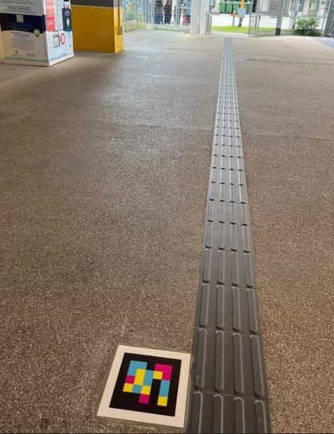 Pasillo exterior del interchange con suelo de granito y una larga línea guía podotáctil gris que se aleja en perspectiva; al inicio, en el suelo, una baldosa cuadrada blanca con el código NaviLens en cuadros amarillos, rosas, azules y negros; al fondo se ven máquinas expendedoras y postes amarillos