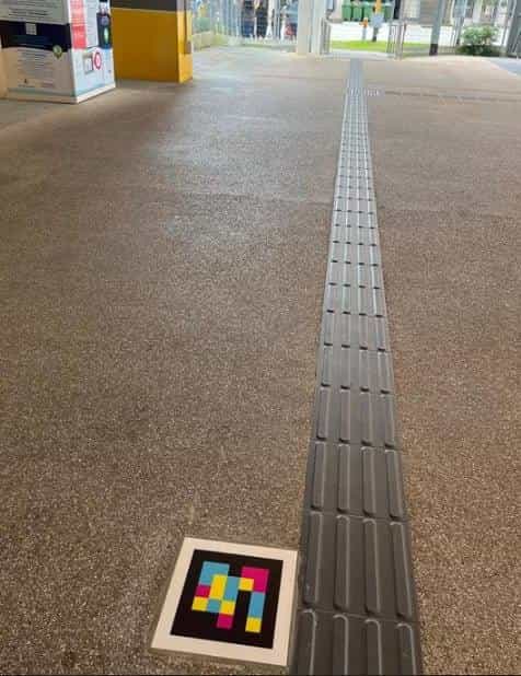 Outdoor walkway of the interchange with a granite floor and a long grey tactile guide line receding in perspective; at its start, on the floor, a square white tile with the NaviLens code in yellow, pink, blue and black squares; in the background, vending machines and yellow bollards